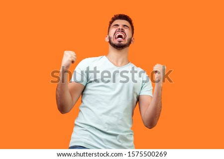 Bottom view of ecstatic motivated brunette man with beard in white t-shirt standing with raised fists and shouting for joy, winner excited for success. indoor studio shot isolated on orange background Royalty-Free Stock Photo #1575500269