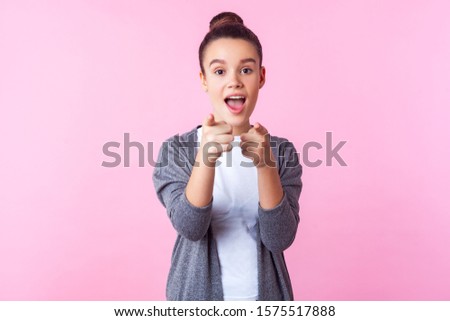 Hey you! Portrait of surprised brunette teenage girl with bun hairstyle in casual clothes pointing at camera, standing with open mouth, wondered astonished expression. studio shot, pink background