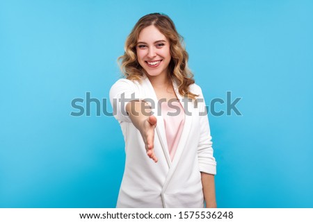 Welcome! Portrait of friendly hospitable cheerful woman with wavy hair in white jacket giving hand to handshake, hostess greeting guests, looking sociable positive. indoor studio shot, blue background Royalty-Free Stock Photo #1575536248