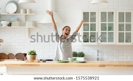 Overjoyed happy young woman raise hands feel excited cooking alone in modern kitchen interior, funny independent girl dancing prepare dinner at home having fun enjoy freedom healthy lifestyle concept Royalty-Free Stock Photo #1575538921