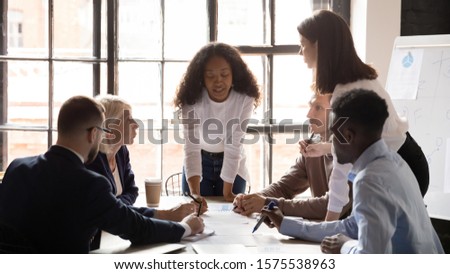 Young african american female professional leaning over table, explaining marketing data analysis paper report to concentrated successful mixed race coworkers at brainstorming meeting in office.