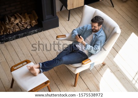 Relaxed casual guy lounge sit on comfortable armchair in modern house room with wooden floor fireplace using smartphone apps, happy young man hold cell phone play mobile games at cozy home, top view