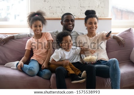 Happy african America parents sit on comfortable couch in living room with little kids watch TV eat popcorn together, smiling biracial family with children enjoy home weekend enjoying movie