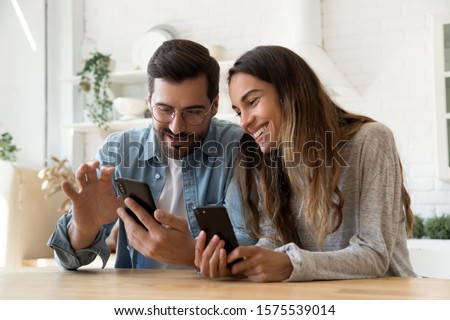 Young happy couple using two phones share social media news at home, smiling husband and wife millennial users customers talking doing shopping online sit at table, mobile tech lifestyle concept