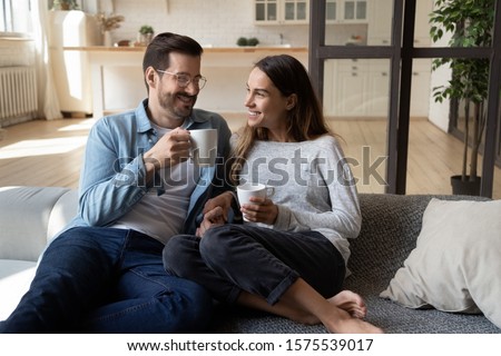 Happy young couple husband and wife relaxing on sofa in modern home living room talking laughing holding cups drinking tea bonding enjoying pleasant conversation romantic leisure lifestyle at home