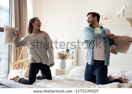 Happy playful young adult romantic couple playing pillow fight on bed together, cheerful carefree man and woman having fun laughing in bedroom interior in morning enjoy lifestyle game at home hotel