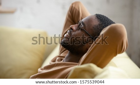 Happy african American young man in glasses sit relax on cozy couch lean hands over head taking nap in living room, smiling biracial male rest on sofa at home sleeping or dreaming, stress free concept Royalty-Free Stock Photo #1575539044