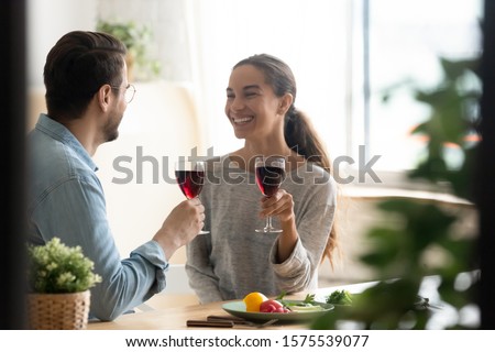 Smiling couple talking laughing celebrating anniversary on date holding glasses drinking red wine, happy husband and wife enjoying healthy food vegetable salad sit at home table on romantic dinner