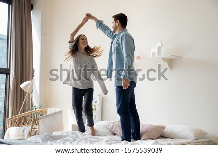 Carefree romantic young couple having fun dancing on bed at modern home, happy active husband and wife enjoying funny party on honeymoon together on leisure morning lifestyle in bedroom interior