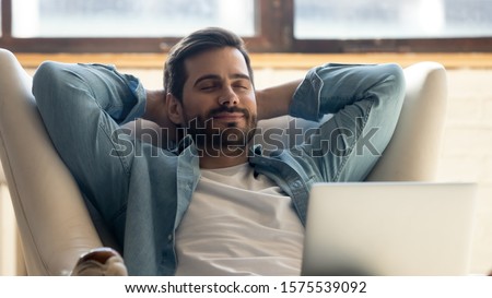 Serene happy healthy young man relaxing on comfortable armchair with laptop, smiling calm relaxed guy lounge eyes closed in sunny cozy home with notebook device enjoying lazy leisure lifestyle on Royalty-Free Stock Photo #1575539092