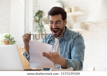 Excited euphoric happy young man holding reading paper postal mail letter amazed overjoyed by good news, got new job celebrate taxes refund receive salary payment loan approval sit at home table Royalty-Free Stock Photo #1575539104