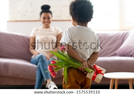 Cute little biracial toddler boy kid stand hiding flowers bouquet and gift making birthday surprise for young mother, small african American child son give present congratulate mom with anniversary