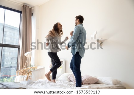 Happy carefree playful young couple dancing on bed together, funny active husband and wife having fun laughing enjoy honeymoon party together in morning in cozy bedroom interior in modern apartment