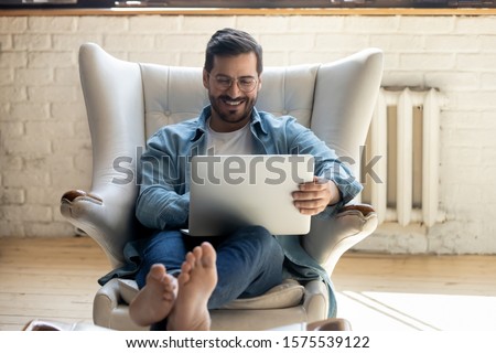 Smiling relaxed young man sit on comfortable armchair using laptop notebook relaxing laughing watching funny movie, happy guy enjoy leisure time indoor with computer having fun online in social media