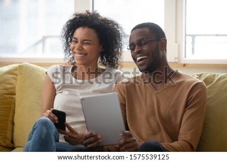 Excited african American couple sit on couch at home relax having fun using modern gadgets together, overjoyed biracial husband and wife rest in living room laugh watching funny video on tablet