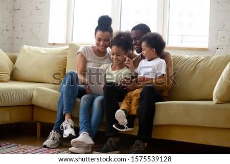 Overjoyed african American young family with little kids sit relax on couch at home watching funny video on tablet, happy biracial parents enjoy weekend with small children using modern pad device