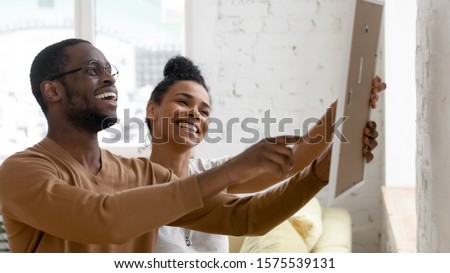 Excited african American young husband and wife hang frame picture on wall happy settling together moving to new house, overjoyed biracial couple decorating own home relocating, ownership concept