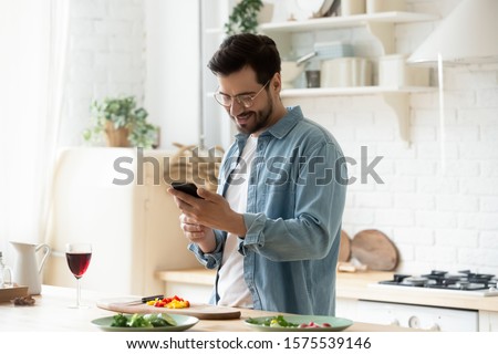 Happy young man preparing romantic dinner searching vegetable recipes diet menu cookbook app using smartphone, smiling husband holding phone cooking healthy vegan food cut salad in kitchen interior Royalty-Free Stock Photo #1575539146