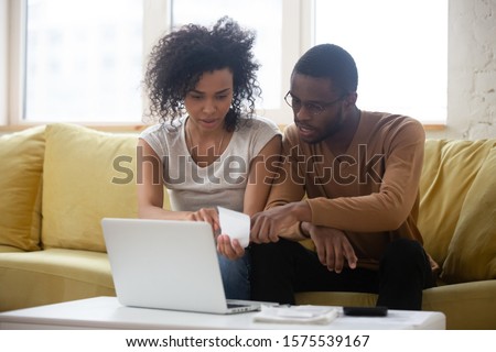 Focused biracial husband and wife sit on sofa in living room pay bills on laptop online managing family finances together, concentrated multiethnic couple busy calculate taxes or expenditures at home
