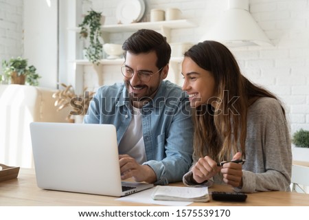 Happy young couple husband and wife using laptop computer looking at screen pay bills online in app calculate mortgage investment payment on website planning budget discuss finances sit at home table Royalty-Free Stock Photo #1575539170