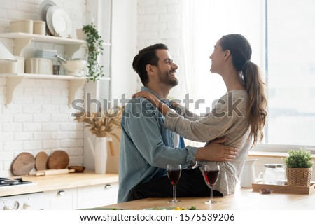 Happy young couple drinking red wine talking bonding in modern kitchen, smiling affectionate husband and wife laughing celebrating romantic anniversary together embracing enjoy married life at home