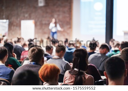 Adult people at conference listen to woman speaker providing lecture on scene in big conference hall. Business and Entrepreneurship concept. Audience at the conference hall. Royalty-Free Stock Photo #1575559552