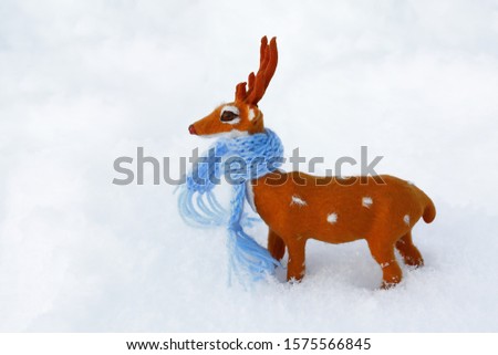 A toy deer in a blue scarf stands among a snowy field. Close up. Christmas picture. Free space on the right. Deer stands sideways to the camera. Shooting from above.