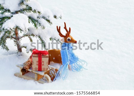 Toy deer in a blue scarf and wooden sled with a gift box under the Christmas tree on a background of a snowy field. Close up, shooting from above. White background. Christmas picture.
