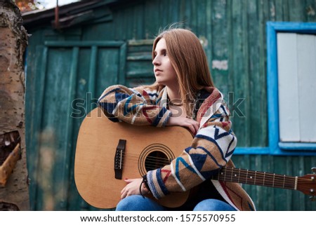 Young blond woman, wearing colorful cardigan, holding acoustic guitar, sitting on a bench in front of old green wooden hut in the woods in autumn. Three-quarter portrait of hippie musician in village