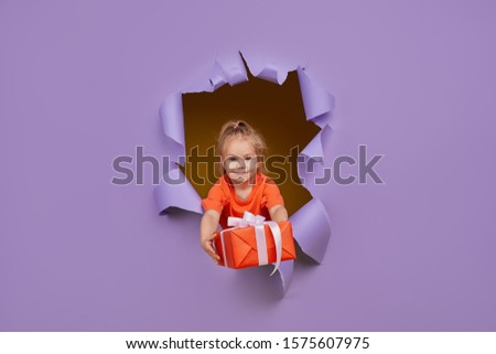 Cute little child girl breaks through a colored purple paper wall with gift box in hands. Toddler funny emotions face. Copy space for text