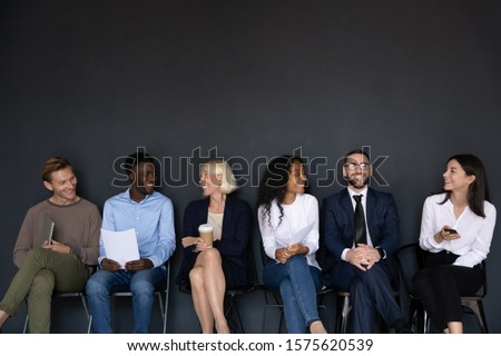 Happy different generations mixed race business people sitting on chairs in row line, isolated on black background with above copyspace. Smiling diverse job seekers chatting, waiting for interview. Royalty-Free Stock Photo #1575620539