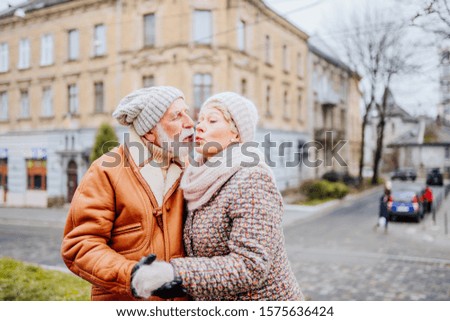 Tourism and technology. Traveling senior couple wearing warm clothes taking a selfie together kissing against the background of attractions of old city street in cold winter outdoor.