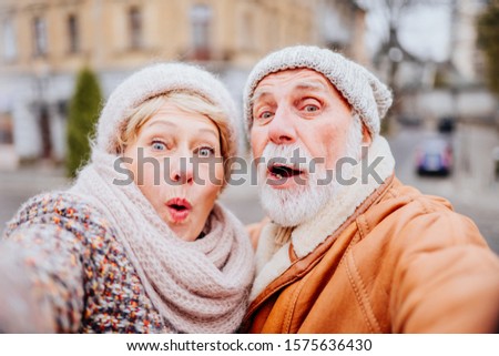 Self-portrait of senior couple is making funny faces crazy cheerful cheery positive playful childish old people life partners having fun on attractions of old city street in cold winter outdoor...