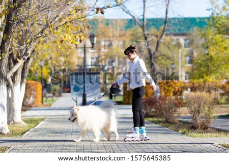 A girl rides on roller skates holding her white dog by leash. Girl learns to skate walking with a dog.