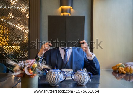 Man sitting in restaurant in suit holds book on level heads. A man in a suit holding a plate instead of his head.