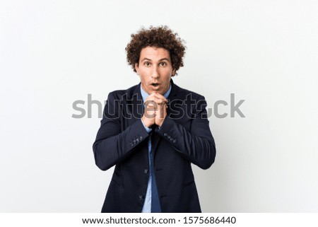 Young business curly man against white background praying for luck, amazed and opening mouth looking to front.