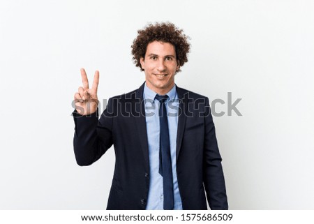 Young business curly man against white background showing victory sign and smiling broadly.