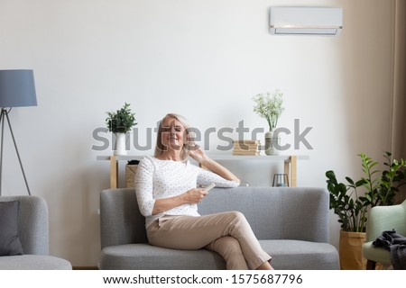 50s woman rest on couch closed eyes enjoy fresh air hold remote control use air conditioner cools herself at summer hot day adjusting temperature inside of living room, comfort wellbeing life concept Royalty-Free Stock Photo #1575687796