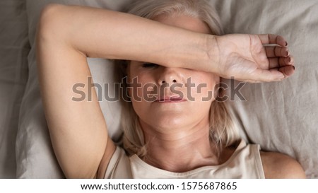 Close up top view middle-aged woman lying down in bed on pillow put hand on face, concept of female having insomnia sleeping disorder or migraine pain, melancholic mood, personal life troubles concept Royalty-Free Stock Photo #1575687865