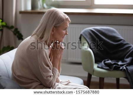 Middle-aged 50s hunched woman seated on couch at home feels upset thinking. Concept of older generation chronic diseases, widow female goes through death of husband, dementia mental disorder concept Royalty-Free Stock Photo #1575687880