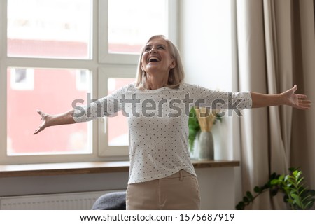 Enjoy of life concept. Lively 60s aged woman standing in warm light living room stretched hands feels very happy wants hug the whole world. Open-hearted female dancing listens music spend time at home Royalty-Free Stock Photo #1575687919