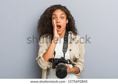 Young african american photographer woman holding a camera shouting excited to front.