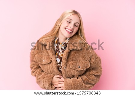 Young blonde woman wearing a coat against a pink background laughs happily and has fun keeping hands on stomach.