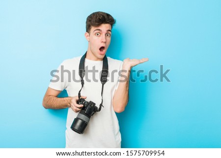Young caucasian photographer man holding a camera impressed holding copy space on palm.
