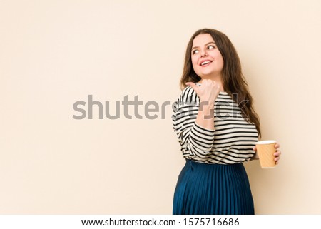 Young curvy woman holding a coffee points with thumb finger away, laughing and carefree.