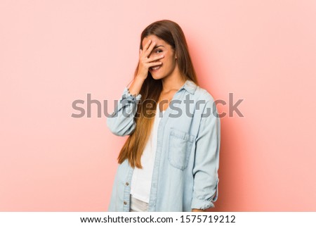 Young slim woman blink at the camera through fingers, embarrassed covering face.