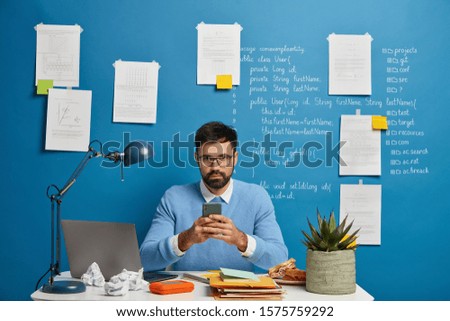 Digital technology expert or enthusiast obsessed with his work, uses mobile phone, works with modern devices, surrounded by many papers, poses at desktop, ready to solve various technical problems.