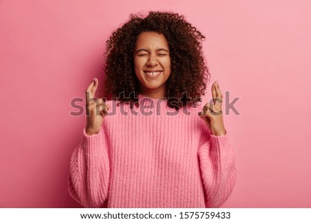 Believing dreams come true. Hopeful dark skinned woman feels lucky, crosses fingers, awaits for miracle, makes wish and has high hopes, smiles broadly, wears oversized sweater, poses indoor.