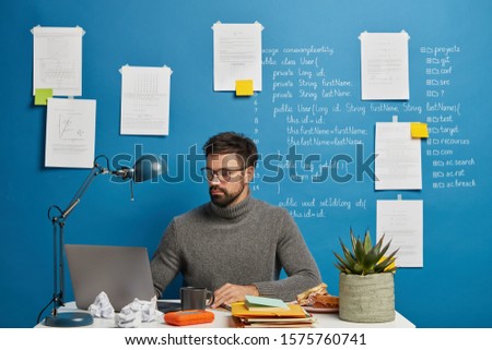 Serious professional male geek concentrated at monitor of modern laptop, wears optical glasses, poses in coworking space against blue background with sticky notes. Programmer works at home office