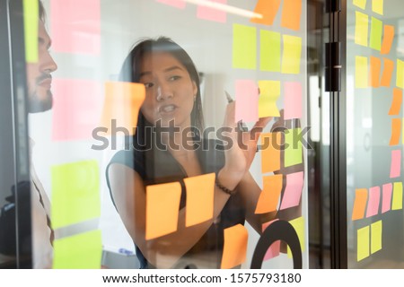 View through glass partition asian female worker working strategizing with caucasian male colleague, attached colourful post-it notes on glass wall where written ideas actions plan for aim achievement Royalty-Free Stock Photo #1575793180